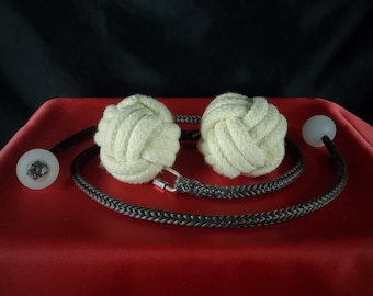 May include: Two cream-colored, knotted rope balls connected by a black and gray braided cord. The cord has a silver clasp and is attached to clear, round, and white objects on each end. The objects are on a red surface with a black background.