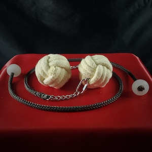 May include: A pair of off-white, knotted balls connected by a black braided rope and a silver chain. The rope has clear, spherical end pieces. The items rest on a red surface against a black background.