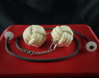 May include: A pair of off-white, knotted balls connected by a black braided rope and a silver chain. The rope has clear, spherical end pieces. The items rest on a red surface against a black background.