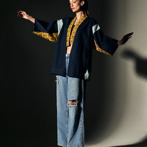 Könnte beinhalten: Dunkelblaue Kimono-Jacke mit gelben und weißen Musterelementen. Dazu werden hellblaue Jeans mit Rissen und ein gelbes Crop-Top getragen. Die Person trägt braune Stiefeletten.