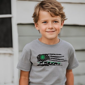 May include: A young boy wearing a gray t-shirt with a distressed American flag design. The flag has a green four-leaf clover in the top left corner and a line of farm animals across the bottom.
