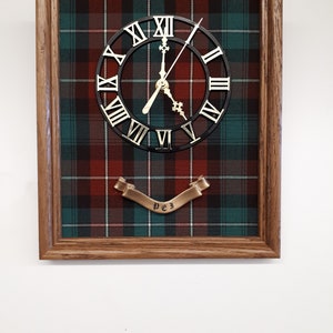 May include: A framed clock with a black face and Roman numerals. The clock is set against a red, green and white tartan background. A gold banner with the word "PEJ" is below the clock.