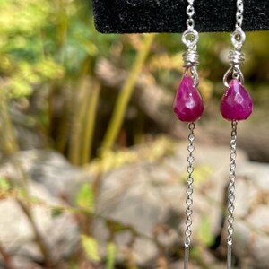 May include: Dangle earrings featuring faceted, teardrop-shaped, deep pink gemstones. The gemstones are suspended from delicate silver chains and wires. The background is a blurred view of greenery and rocks, creating a natural setting for the jewelry.