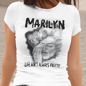 May include: White t-shirt featuring a black and white sketch of Marilyn Monroe with her hand covering her eyes. The word "MARILYN" is printed above the image, and the phrase "LIFE AIN'T ALWAYS PRETTY" is below.