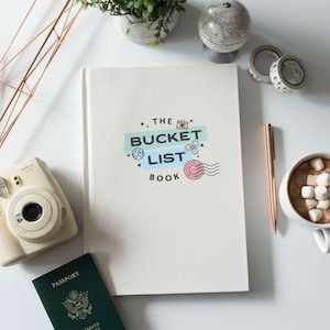 May include: A white hardcover book titled THE BUCKET LIST BOOK with travel-themed icons including a camera, globe, and postmark. The book is surrounded by a cream-colored instant camera, a dark green passport, a rose gold pen, and a mug of hot cocoa.