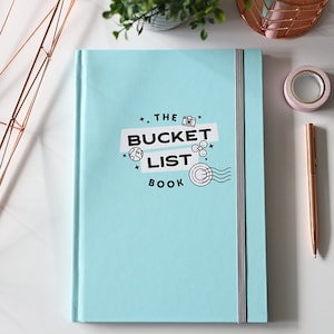 May include: A light blue journal with a white elastic closure. The cover has a black and white graphic design that says "The Bucket List Book" with a camera, globe, and other travel-related icons.
