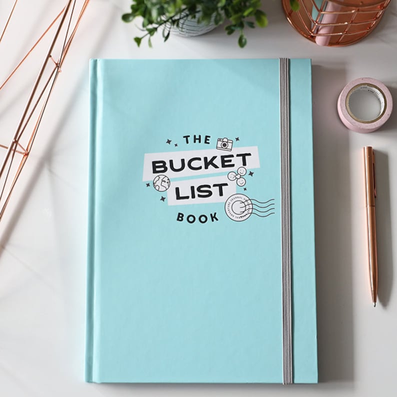 May include: A light blue journal with a white elastic closure. The cover has a black and white graphic design that says "The Bucket List Book" with a camera, globe, and other travel-related icons.