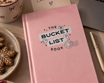 May include: A light pink hardcover book titled The Bucket List Book with black text, a camera icon, a globe, smiley faces, and a postmark stamp graphic on the front cover. The book is positioned on a surface next to a gold pen and a note that reads 100 days till Christmas.