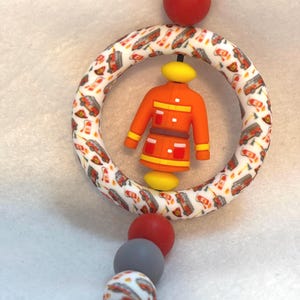 May include: A baby teether featuring a firefighter theme. The design includes a ring with a pattern of fire trucks, a small orange firefighter figure, and a strand of colorful beads in red, gray, and white.