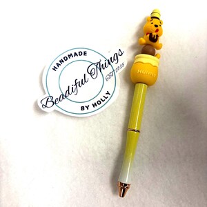 May include: A yellow pen with a Winnie the Pooh design. The pen features a yellow barrel with a white and gold tip. The pen has a Winnie the Pooh figure on top of a honey pot. The pen has the word "Hunny" on the honey pot.