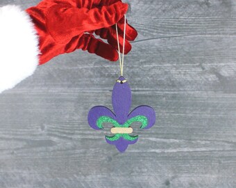 May include: A purple fleur-de-lis ornament with green glitter accents and a gold string for hanging. The ornament is held by a person wearing a red velvet glove with white trim. The background is a gray wooden surface.