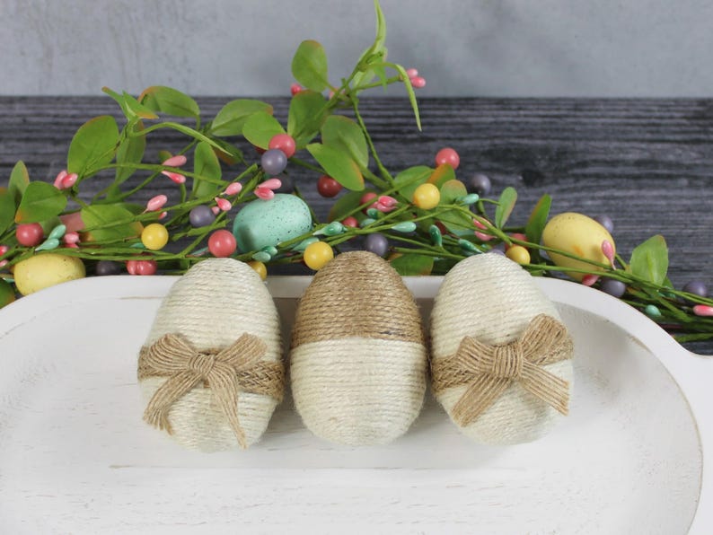 以下が含まれることがあります： 茶色と白のひもで包まれ、茶色のリボンで飾られたイースターエッグが3つ。白いボウルに入っています。