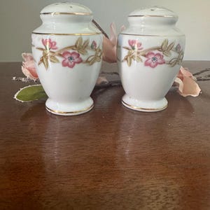 May include: A set of two white porcelain salt and pepper shakers with a floral design. The shakers have a gold trim around the top and bottom.