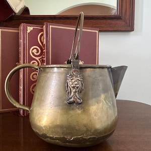 May include: Antique brass teapot with a lion head design on the front. The teapot has a handle and a spout. It is sitting on a wooden table.