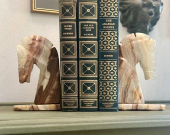 May include: A set of three antique books with gold lettering and dark green covers, held between two marble horse head bookends. The books are titled "Oliver Twist", "The Old Curiosity Shop", and "The Arabian Nights".