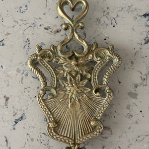 May include: A gold-toned metal hot pad with an ornate design. The pad features a starburst pattern in the center and a heart-shaped top.