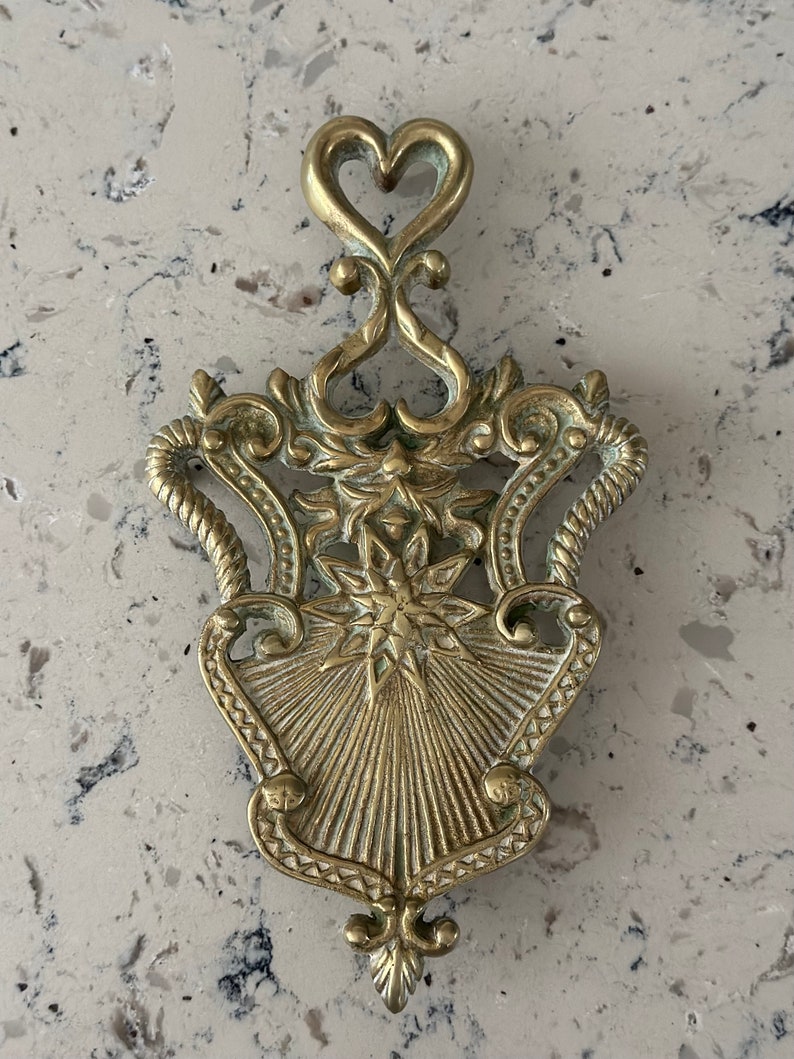 May include: A gold-toned metal hot pad with an ornate design. The pad features a starburst pattern in the center and a heart-shaped top.