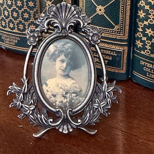 May include: An antique silver-toned oval picture frame with a portrait of a woman. The frame has floral and shell-like decorative elements. The background includes several antique books with gold lettering and dark green covers.
