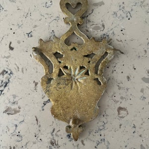 May include: A gold-colored metal hot pad with an intricate design featuring a heart shape at the top and a fleur-de-lis at the bottom.