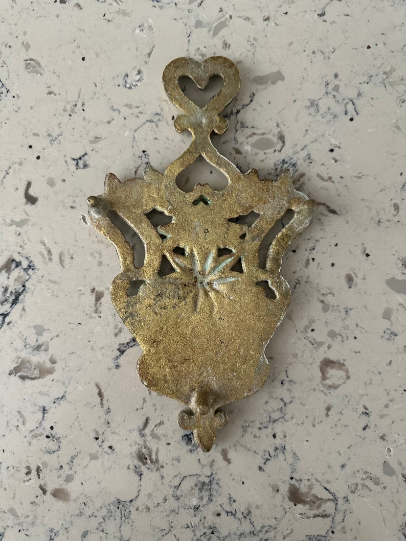 May include: A gold-colored metal hot pad with an intricate design featuring a heart shape at the top and a fleur-de-lis at the bottom.