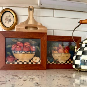 May include: Two rectangular wooden frames containing artwork of fruit in bowls. The left frame shows red apples in a bowl, while the right frame displays a variety of fruits in a bowl. Both images feature a checkered pattern at the base.