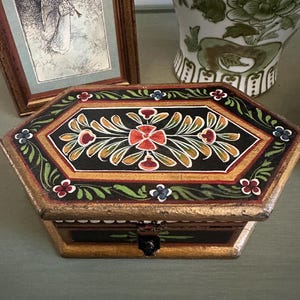 May include: Hexagonal wooden box with a hinged lid. The box is painted black with a floral design in red, orange, green, and blue. The edges are gold-colored, and the box has a small black clasp.