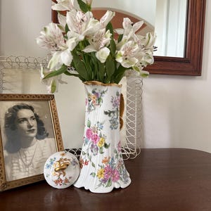 May include: A white porcelain vase with a gold rim and floral design, holding white and pink flowers. A framed vintage photograph and a decorative lid with a gold handle are nearby. The vase is approximately 10 inches tall.