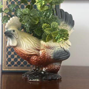 May include: A ceramic rooster planter with a cream, brown, and gray color scheme. The planter is filled with green artificial foliage. The rooster has a detailed design and is placed on a wooden surface.