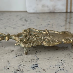 May include: A gold-toned metal trivet with an ornate design. The trivet has three legs and a decorative handle.