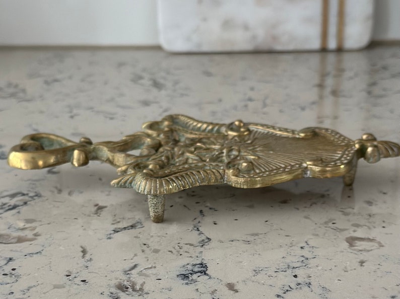 May include: A gold-toned metal trivet with an ornate design. The trivet has three legs and a decorative handle.