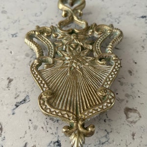 May include: A gold-toned metal hot pad with an ornate design. The pad features a starburst pattern in the center and a heart-shaped top.