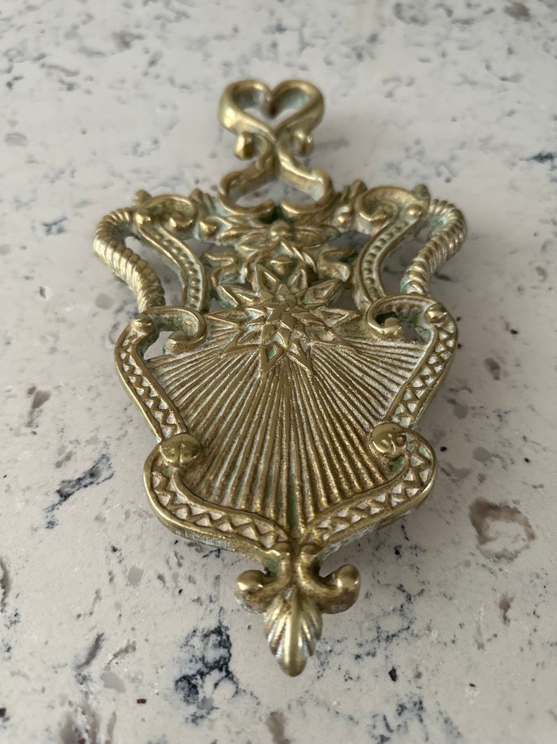 May include: A gold-toned metal hot pad with an ornate design. The pad features a starburst pattern in the center and a heart-shaped top.