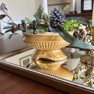 May include: A yellow ceramic bowl with a decorative base sits on a mirrored tray. The bowl is filled with faux greenery, berries, and a pine cone.