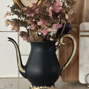 May include: A black teapot-shaped vase with gold accents, holding dried pink flowers. The vase has a decorative handle and sits on a gold-colored base. A framed picture is in the background.