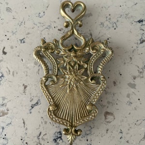 May include: A gold-toned metal trivet with an ornate design featuring a heart at the top and a starburst pattern in the center.