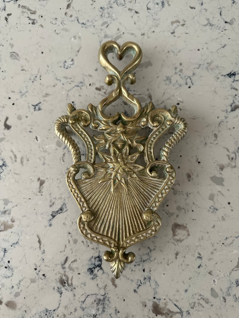 May include: A gold-toned metal trivet with an ornate design featuring a heart at the top and a starburst pattern in the center.