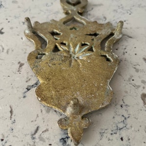 May include: A gold-toned metal hot pad with an ornate design. The pad has a heart-shaped top and a decorative bottom.