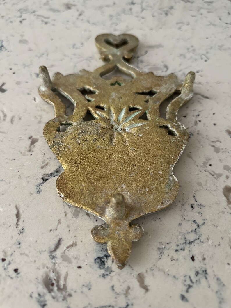 May include: A gold-toned metal hot pad with an ornate design. The pad has a heart-shaped top and a decorative bottom.
