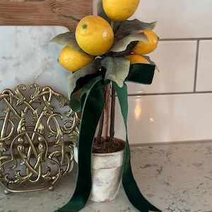 May include: A decorative lemon topiary in a distressed white pot, adorned with bright yellow lemons, green leaves, and a dark green velvet ribbon. The artificial plant adds a touch of freshness to any space.