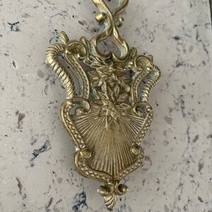 May include: A gold-toned metal trivet with an ornate design. The trivet has a heart-shaped top and a decorative, sunburst-like center.
