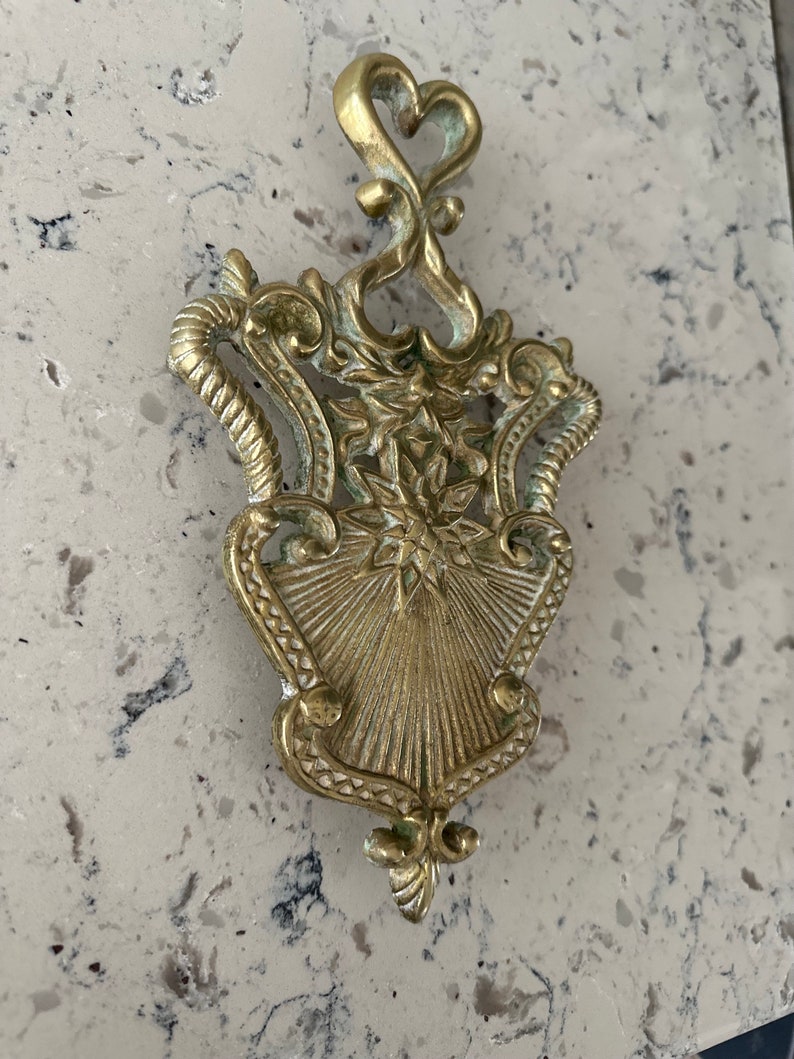 May include: A gold-toned metal trivet with an ornate design. The trivet has a heart-shaped top and a decorative, sunburst-like center.