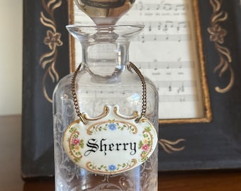 May include: A clear glass bottle with a decorative stopper and a porcelain tag that reads "Sherry". The tag is decorated with floral designs in pink, blue, and yellow.