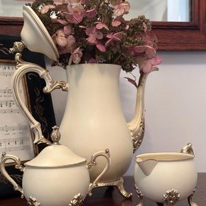 May include: A cream-colored tea set with gold accents, including a teapot with dried flowers, a sugar bowl, and a creamer. The set has ornate handles and decorative feet, creating an elegant, vintage aesthetic.