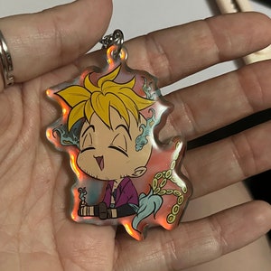 May include: A holographic keychain with a cartoon character design. The character has blond hair and a purple shirt. The keychain has a silver chain and a colourful, iridescent design with orange and blue accents.