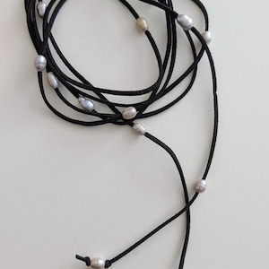 May include: A black cord necklace with several oval-shaped pearls. The necklace is looped and knotted, with the pearls spaced along the cord. The pearls are a mix of white and grey. The necklace is on a white surface.