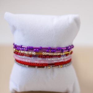May include: A stack of six beaded bracelets featuring a variety of colors including purple, gold, red, light pink, and silver. The bracelets are constructed from small cylindrical and round glass beads arranged in alternating patterns on each strand.
