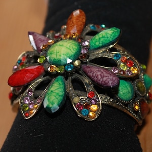 Peut inclure: Un bracelet en forme de fleur coloré avec des pierres vertes, rouges, violettes et orange. Le bracelet est en métal et a un aspect vintage.
