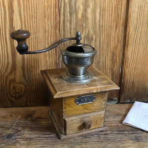 May include: Antique wooden coffee grinder with a metal crank and a drawer. The grinder has a label that reads "De Luxe".