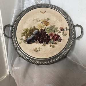 Puede incluir: Una bandeja de servicio redonda de metal plateado con un borde trenzado estilo cuerda y dos asas ornamentadas. El centro presenta una incrustación circular que muestra una ilustración de bodegón con uvas verdes, ciruelas y flores.