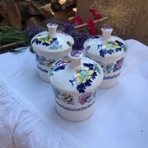 Op de afbeelding: Drie witte keramische kruidenpotten met deksels. De potten hebben een bloemenpatroon in blauw, geel, roze en groen. De deksels hebben een klein tuitje om te gieten.